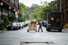 In this May 22, 2020, photo, a delivery man pushes a cart full of packages to deliver to an apartment building on an almost…