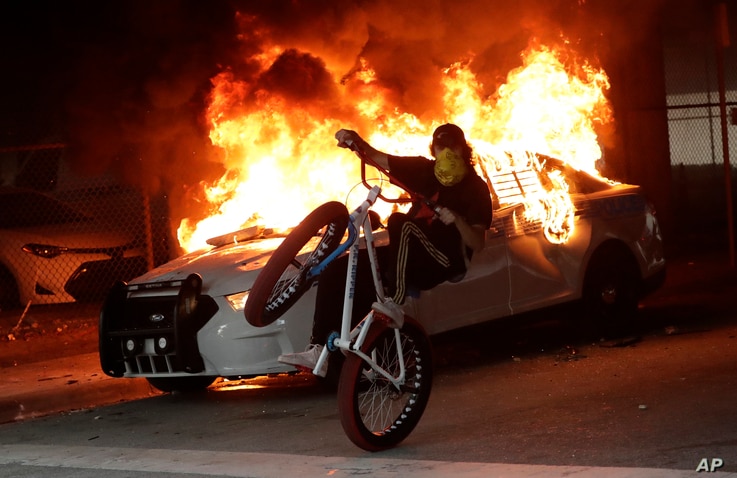 A protester on a bicycle rides past a burning police car during a demonstration next to the city of Miami Police Department,…